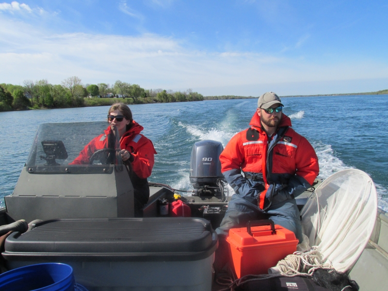 Two people sit at the back of a boat while one drives. There is a net next to one of them, and both are wearing safety suits.