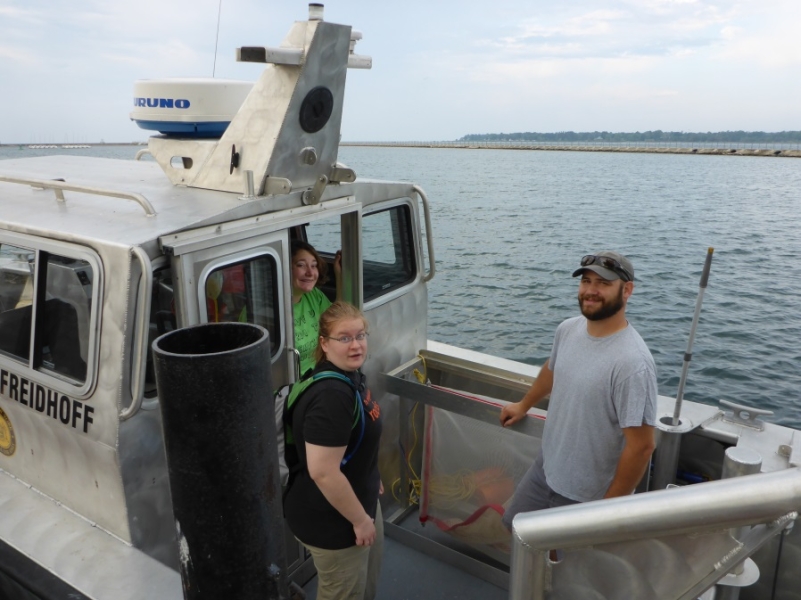 Three people stand on the back of a boat with a cabin.