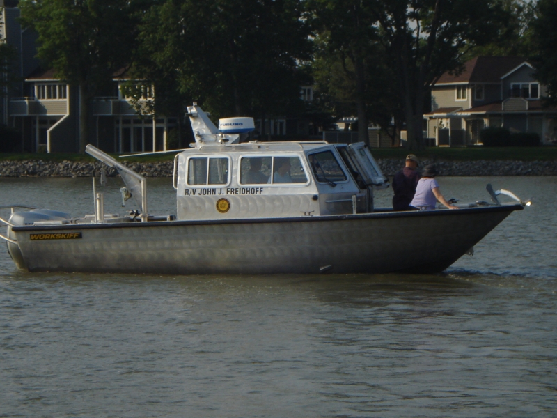 Two people stand in the front of a boat with a cabin named "R/V John J. Freidhoff."