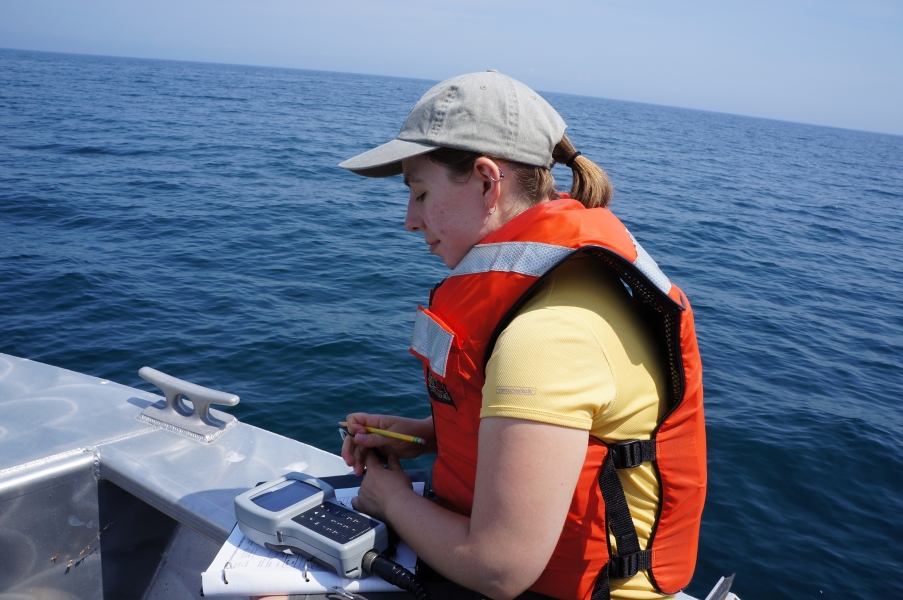 A person in a life jacket sits at the edge of a boat with an instrument, a datasheet, and a pencil