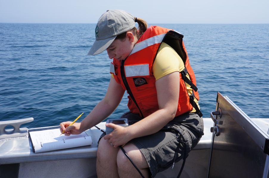 A person in a life jacket sits at the edge of a boat with an instrument in their lap. They are writing on a datasheet.