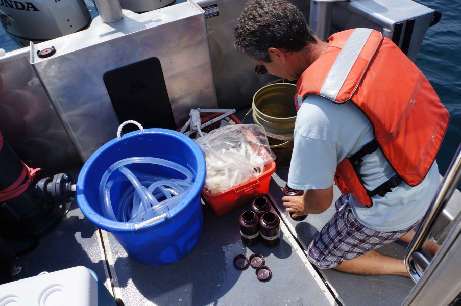 A person in a life jacket lines up bottles on the deck of a boat. There is a tub with a tube in it, and two other buckets nearby.