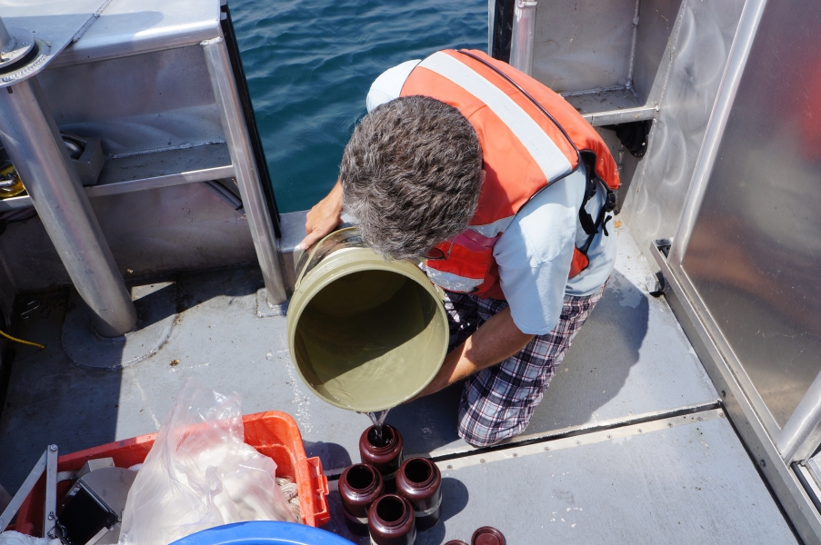 A person in a life jacket pours water from a bucket into bottles lined up on the deck of a boat