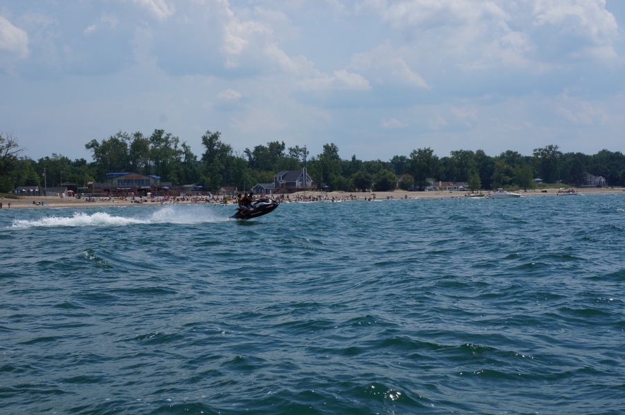 A person rides a jet ski by a beach full of people. There are a few small boats anchored near the beach.