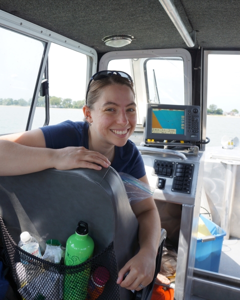 A person smiles, looking over the back of a seat inside the cabin of a boat. There is a screen with a map behind them.