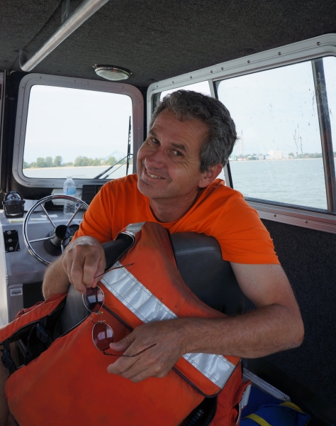 A person smiles, looking over the back of a seat inside the cabin of a boat. They have a life jacket over their seat and are holding sunglasses in their hands.