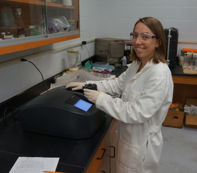 A person wearing a lab coat, safety glasses, and gloves stands next to an instrument with a screen on a counter in a lab, holding a carousel with cuvettes