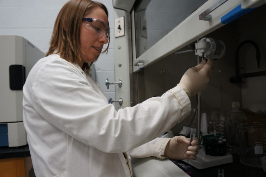 A person in a lab coat concentrates and works at a fume hood, pipetting a liquid from a sample tube into a cuvette