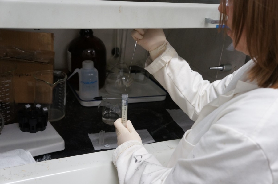 A person in a lab coat works at a fume hood, pipetting a liquid from a sample tube into a cuvette