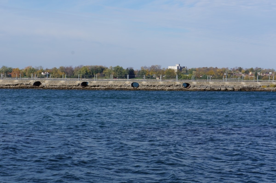 A breakwall that separates the canal from the the river. At least a foot of gravel is visible below the culverts in the breakwall. There is an area of wet gravel above the current water level, as if the water level has fallen.