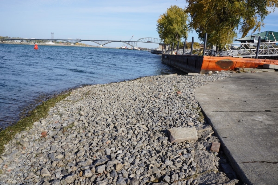 A stretch of gravel leading down to the water, and weeds at the waterline. There is a concrete pad further up the slope from the gravel. A barge is next to the ramp and a bridge in the distance