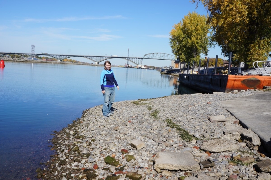 A person stands on the gravel by the waterline. The dock and concrete pad are about ten feet up the ramp. There is a line of weeds washed up about halfway between the water and the concrete pad, marking a previous waterline.