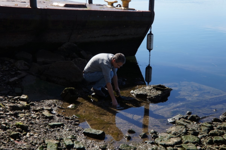 A person crouches on the ground next to the barge dock, putting their hand in very shallow water.