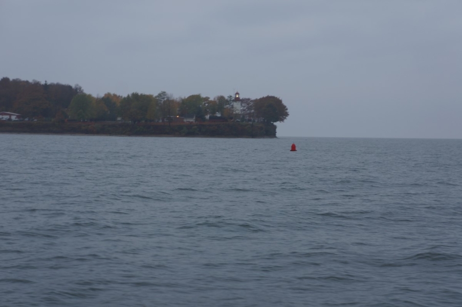 A cliff with trees and a lighthouse, surrounded with water. There is a red navigational buoy in the water.