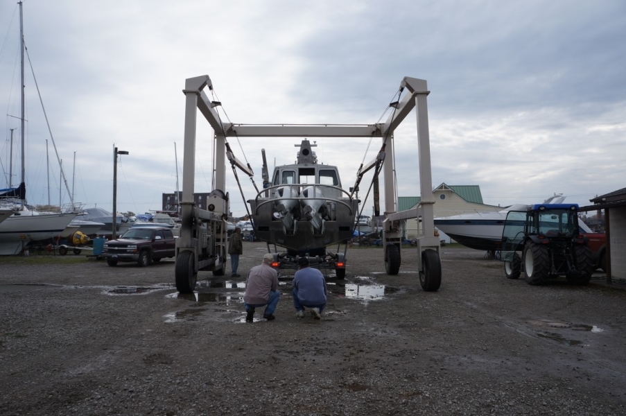 Two people crouch to check that a trailer is lined up under a boat. The boat is held aloft by straps on a metal frame.