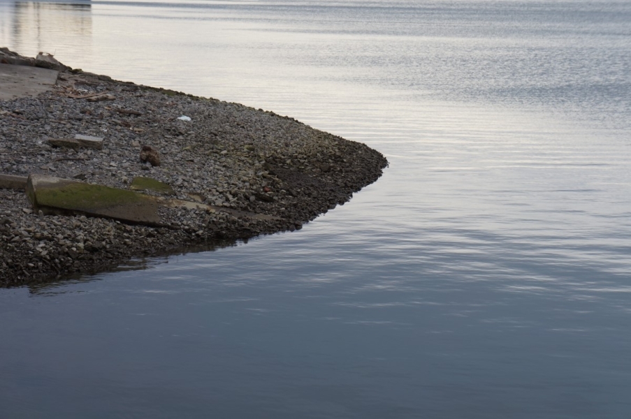 A stretch of gravel leading down to the water, the slope beginning to get steeper. There is a concrete pad further up the slope from the gravel