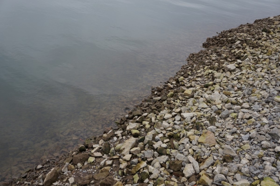 A steep stretch of gravel leading down to the water. Some of the stones are grey while the ones closer to the water are brown, possibly because they are wet or covered in algae, as if they are normally covered.