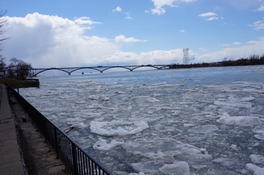 Ice chokes the river near the break wall. There is a bridge in the distance.