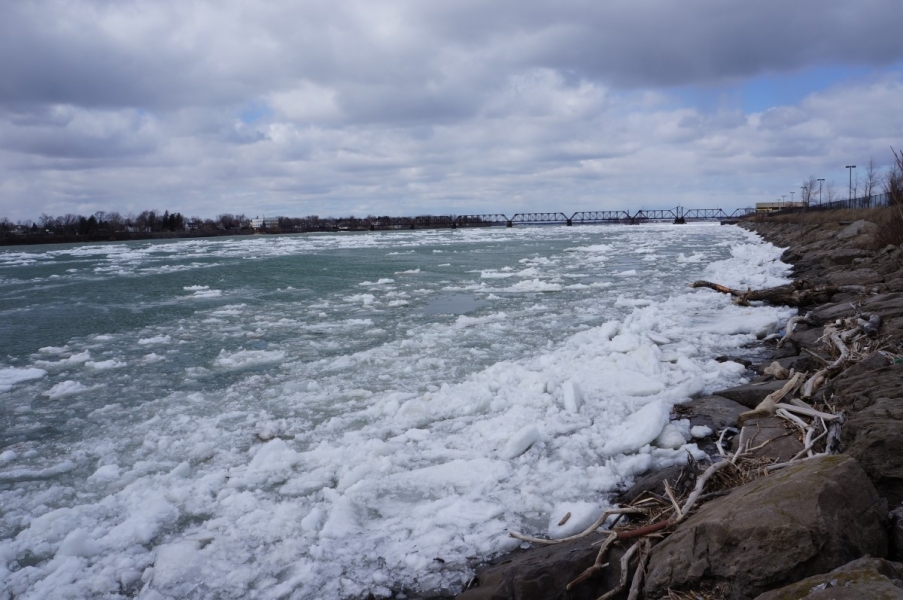 Ice piling up along the shore of the river. There is a train bridge in the distance.