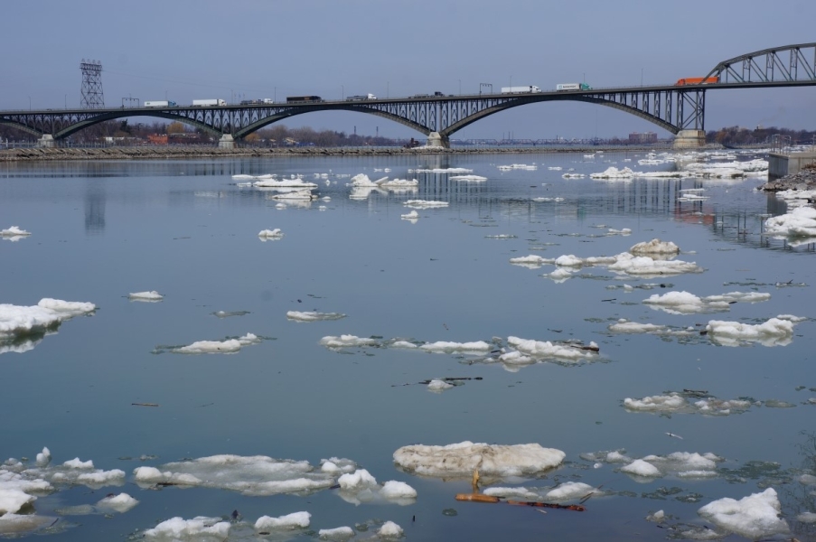 A few pieces of ice in the water. There is a breakwall and a bridge in the distance.