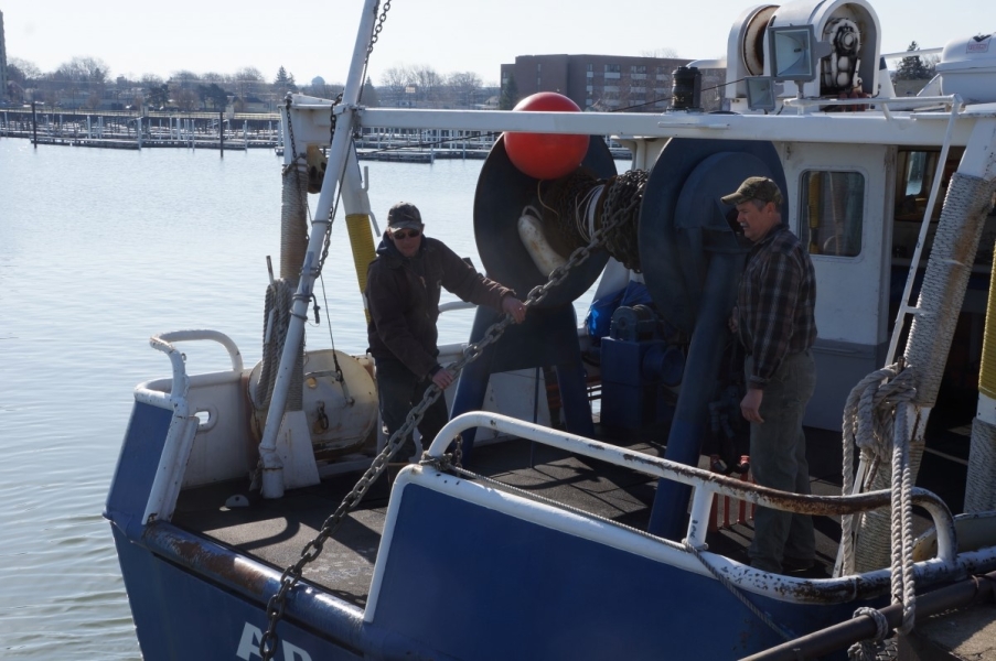 Two people coil chain onto a large reel on the back of a boat.