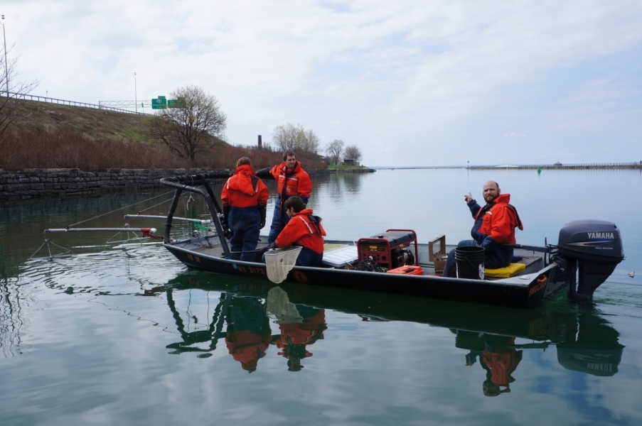 Four people in coldwater safety gear on a shallow boat with a railing at the front and two booms with wires hanging into the water. Two people stand at the front of the boat, one sits with a net, and the last is at the back of the boat by the controls