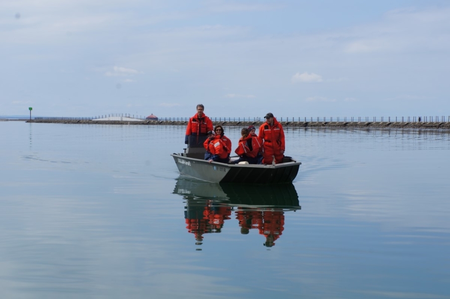 A group of people in coldwater safety gear aboard a shallow boat on glass-calm water