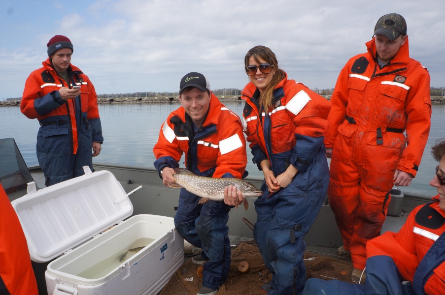 Five people in coldwater safety gear pose for a picture on a boat. One is holding a large brown speckled fish while standing next to a cooler with other fish in water