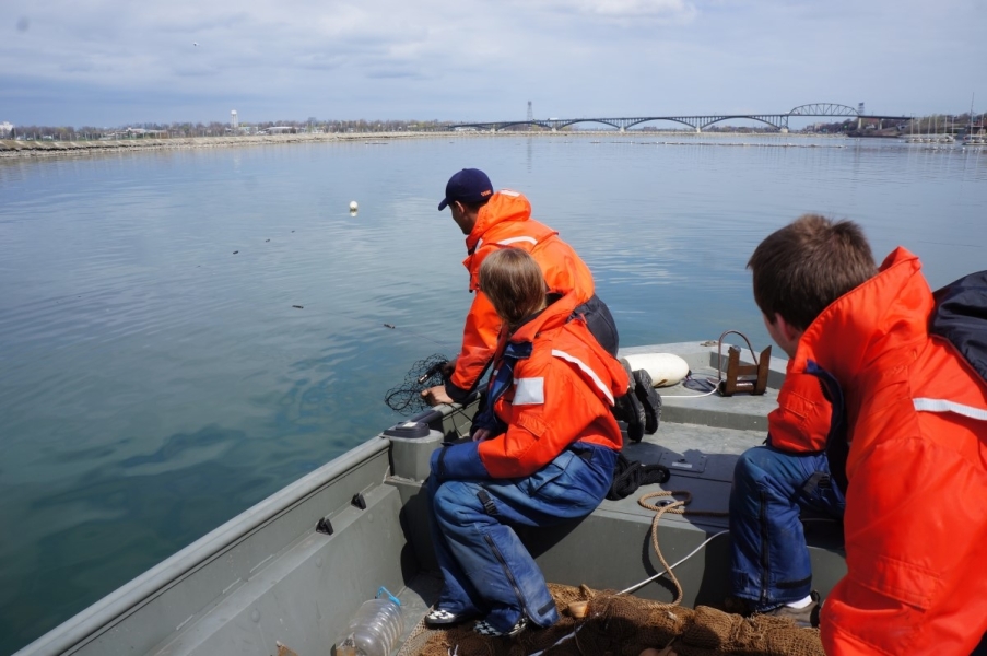 Three people in coldwater safety gear sit at the front of a boat with some nets on it, looking toward some small floats in the water