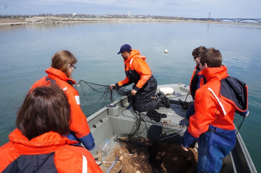 Five people in coldwater safety gear at the front of a boat pulling in a square framed net