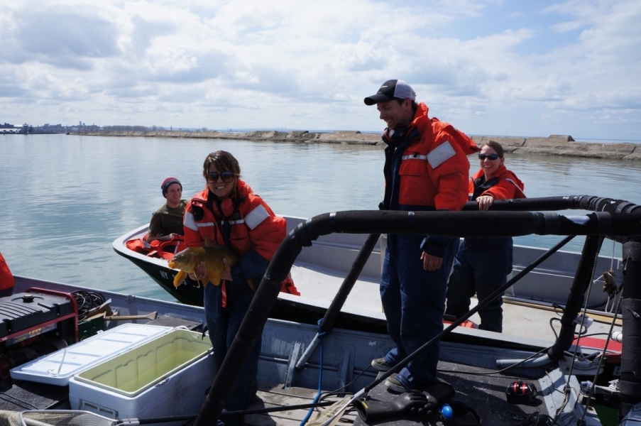 Two boats pulled side by side. People in the farther boat look at the two people in the closer boat who are standing by an open cooler full of water. One person holds a large fish in their arms