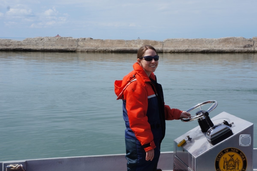 A person standing up at the helm of a boat. A breakwall is the the background