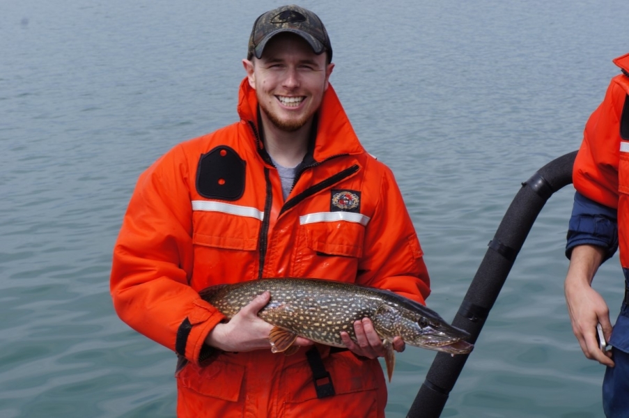 A person smiles and stands in front of water holding up a large speckled fish