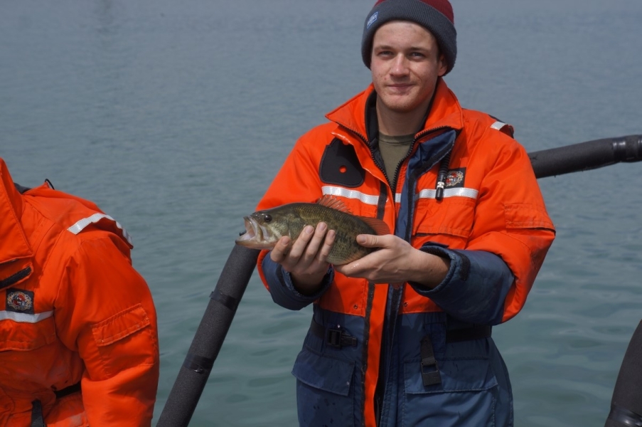 A person stands in front of water holding up a medium fish with a large gaping mouth