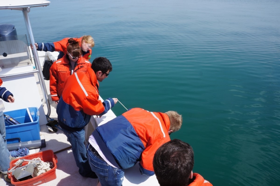 People in coldwater safety gear lean over the side of a boat to watch one of them deploy some equipment