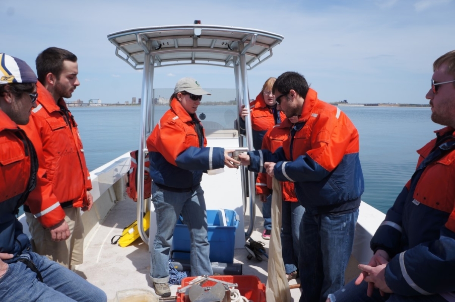 People on a boat looking at equipment. One holds a net while another person hands them a small metal cup