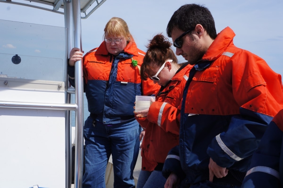 Three people in coldwater safety gear stand on a boat. One looks at water in a clear plastic jar