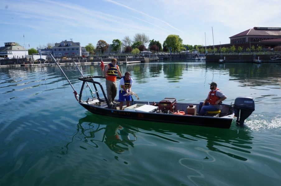 Three people on a shallow boat with a railing. One person operates the motor while two stand in the front. One person is standing over a cooler and holding up a large fish.