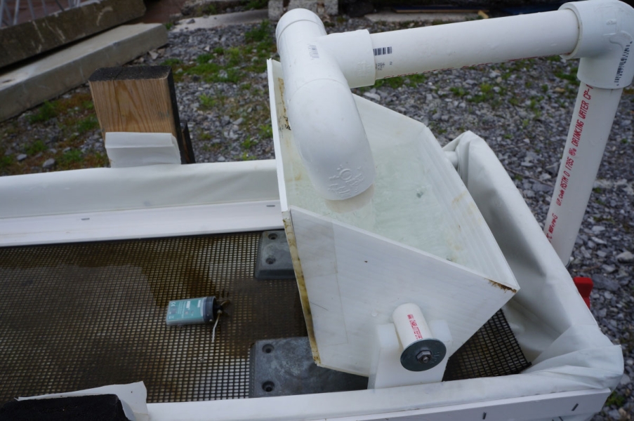 Water flows from a pipe into a small rectangular bucket over a trough with some algae and a small green sensor