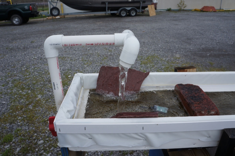 Water flows from a pipe onto a narrow trough with algae, rocks, and a small green sensor