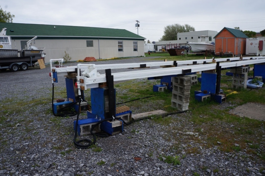 two long thin troughs held up about three feet from the ground with water dripping from a pipe into them, set up in front of a building. A boat on a trailer is partially visible in the background