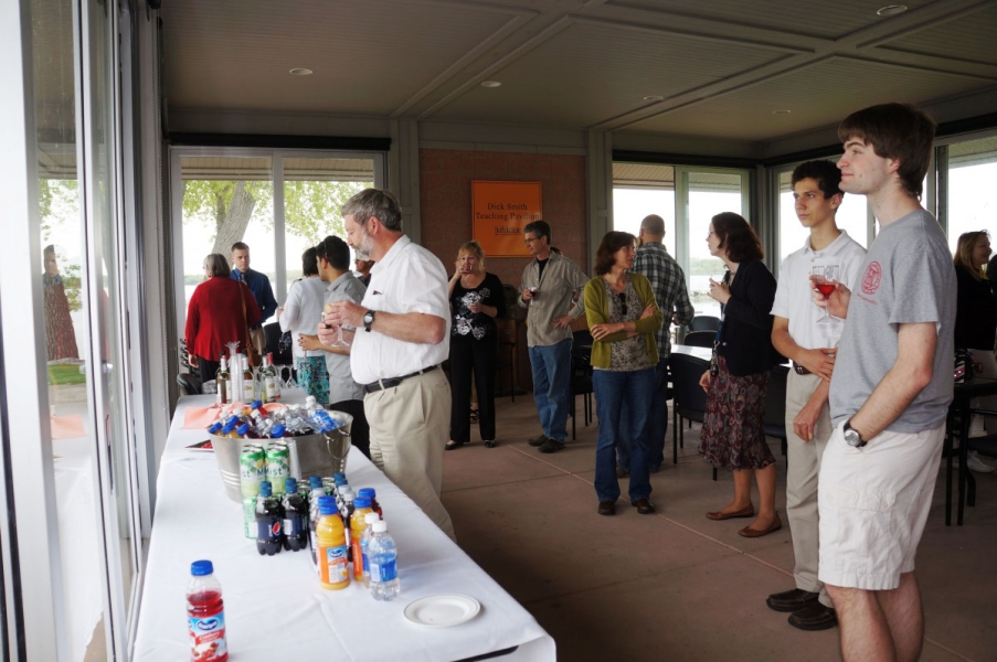 People stand in a pavilion with large glass windows, getting drinks from a table along one wall.