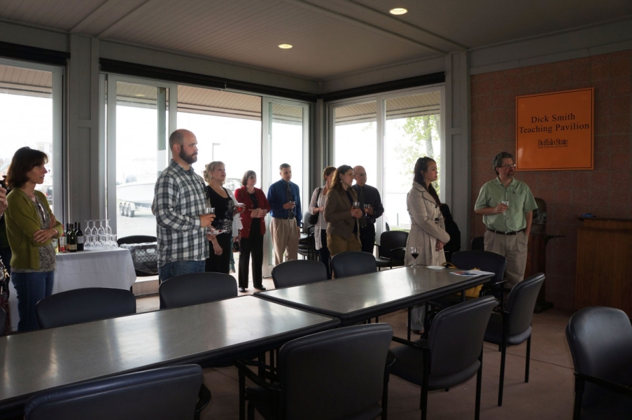 People stand facing the same direction to watch someone speak. They are in a pavilion with glass walls.