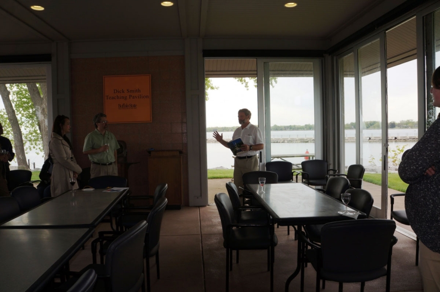 A person speaking to a group of people inside a pavilion with glass walls. A waterway is in the background.