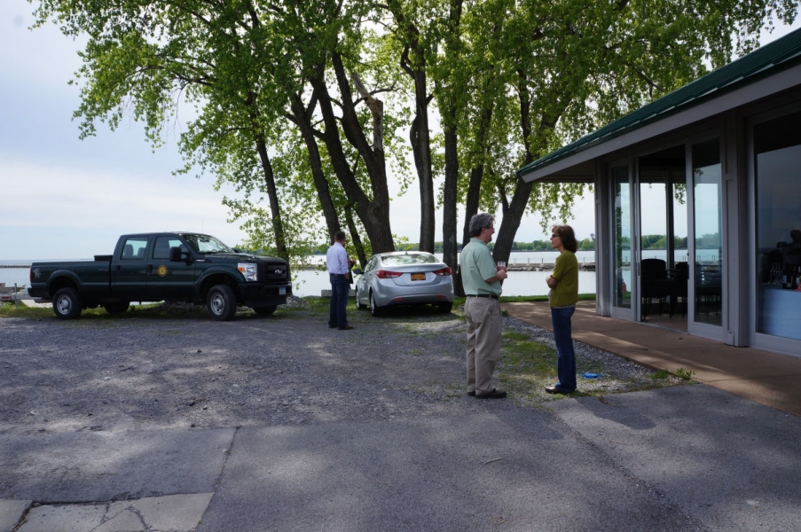 People stand by some vehicles near a pavilion building. Behind them is a tall tree and a waterway.