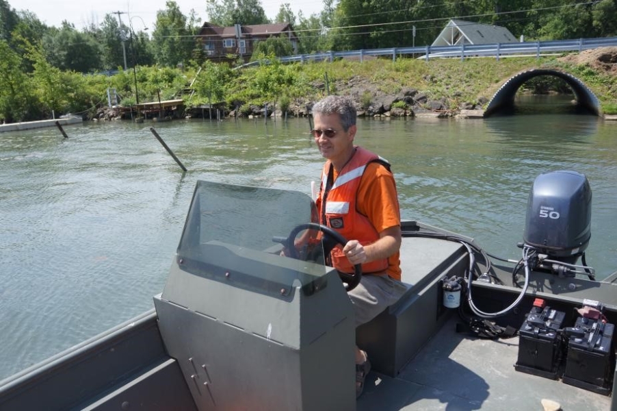 A person sitting at the controls of a boat. The shore is nearby, with a large culvert.