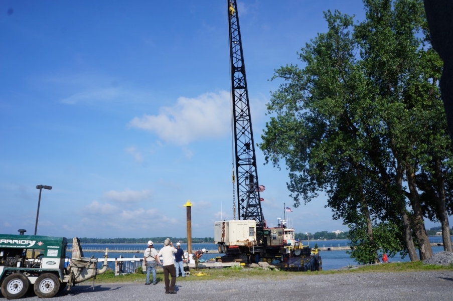 A tug and a barge with a crane approach a dock while people in hard hats wait