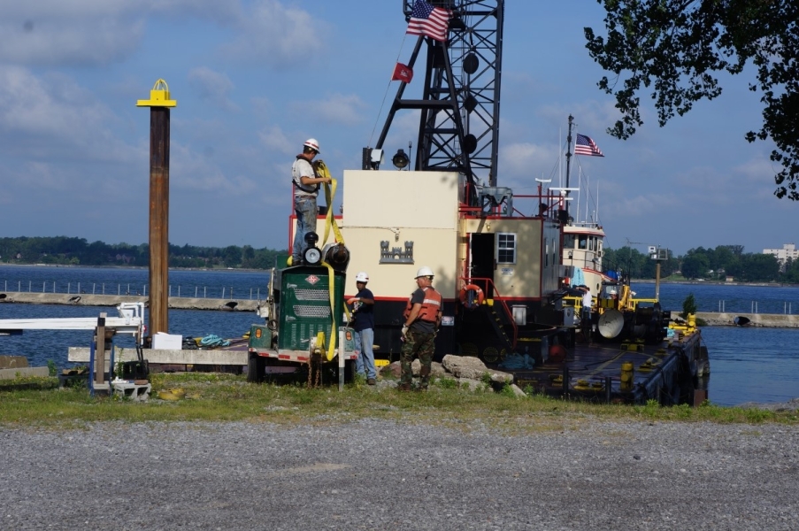 Crew members put straps on a generator while a crane on a barge stands by behind them.