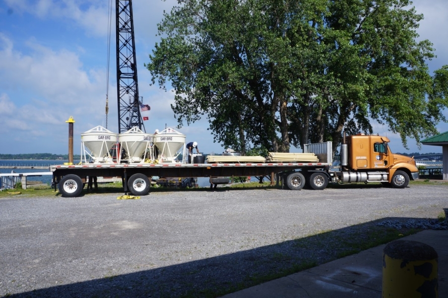 A truck arrives at the waterfront with three concrete vats and some planks. There is a crane behind them.