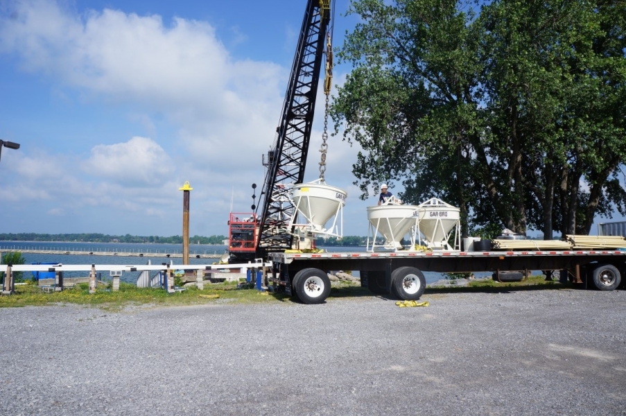 A crane on a barge lifts a concrete vat off the flatbed of a truck. There are two other concrete vats.